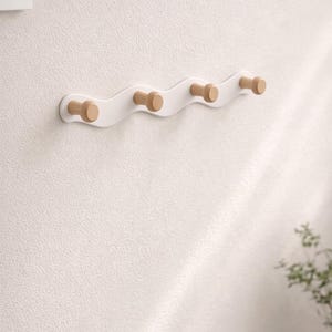 Può includere: Un attaccapanni a parete bianco ondulato con quattro pioli in legno. L'attaccapanni è montato su una parete bianca strutturata. I pioli sono cilindrici e in legno chiaro. Il design è minimalista e moderno.