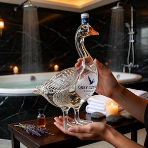 Puede incluir: Una botella de vidrio transparente con forma de ganso, que presenta la etiqueta Grey Goose Vodka France con un logotipo de ganso azul. La botella tiene un cuello largo, detalles de alas texturizados y un pico naranja. Se sostiene frente a un baño.