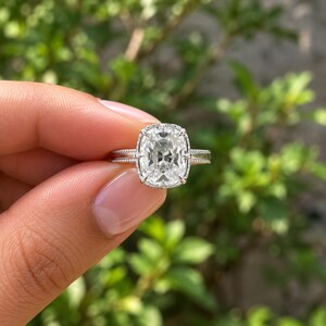 Op de afbeelding: Een zilverkleurige ring met een grote, heldere edelsteen in kussenslijpsel in een klauwzetting. De ringband heeft een verfijnde kralentextuur aan de zijkanten, wat het sieraad een gedetailleerde en elegante uitstraling geeft in het ontwerp.