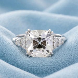 Peut inclure: Une bague en platine avec une grosse pierre précieuse transparente de taille carrée au centre. De plus petites pierres précieuses transparentes flanquent la pierre principale de chaque côté. La bague est posée sur un fond en tissu bleu clair.