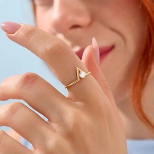 Könnte beinhalten: Ein goldfarbener Ring mit V-förmigem Design und einem birnenförmigen, klaren Edelstein. Der Ring befindet sich an einem Finger, mit einem unscharfen Hintergrund. Das Band des Rings ist dünn und filigran, der Edelstein ist in einer Krappenfassung gefasst.