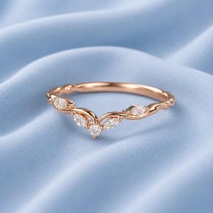 Peut inclure: Bague en or rose avec un design délicat et incurvé. La bague présente plusieurs petites pierres précieuses claires serties le long de la bande, avec un groupe de trois pierres au centre. La bague est posée sur un fond bleu clair.