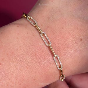 May include: A gold link bracelet with rectangular links, some set with small, sparkling stones. The bracelet is on a wrist against a burgundy background. The brand name "Iveljewels" is visible.