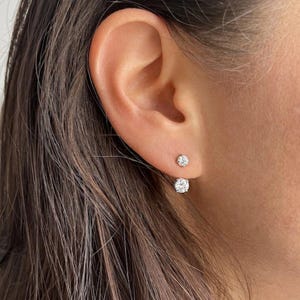 May include: A double-sided earring featuring two round, clear, faceted stones set in silver-toned metal. The smaller stone sits directly on the earlobe, while the larger stone hangs below the lobe, creating a layered, suspended appearance.