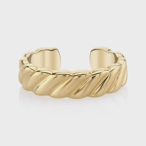 May include: A gold-toned open ring featuring a continuous twisted rope texture along the band. The metal has a polished, reflective finish with a distinct gap at the centre, creating a circular shape with a repeating diagonal pattern throughout.