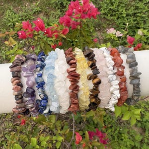 Può includere: Una collezione di braccialetti con schegge di pietre preziose in vari colori, tra cui marrone, viola, blu, bianco, giallo e rosa. I braccialetti sono esposti su una superficie cilindrica bianca, con sfondo di foglie verdi e fiori rosa.