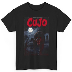 May include: Black t-shirt featuring a graphic from the horror novel "Cujo." The design includes the title in red, a menacing dog with glowing red eyes, a full moon, and a spooky graveyard scene. "Terror Has a New Name" is printed above the title.