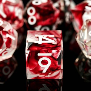 May include: A four-sided square die featuring a translucent resin body with swirling deep red and clear patterns. The die displays white numerical markings, including the number 4 at the top and the number 6 at the bottom, set against a dark background.