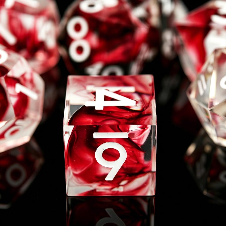 May include: A four-sided square die featuring a translucent resin body with swirling deep red and clear patterns. The die displays white numerical markings, including the number 4 at the top and the number 6 at the bottom, set against a dark background.