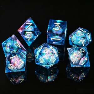 Puede incluir: Juego de siete dados poliédricos con un diseño celestial. Los dados son de color azul transparente con números dorados y contienen inclusiones coloridas y brillantes. Los dados están dispuestos sobre una superficie negra reflectante.