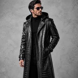 Può includere: Un lungo trench nero con motivo testurizzato in pelle di coccodrillo, cappuccio, ampi revers e polsini abbottonati. Il cappotto è indossato sopra un maglione nero a collo alto, pantaloni neri e stivali stringati, con occhiali da sole.