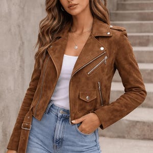 Puede incluir: Chaqueta de ante marrón de estilo motero, con cuello de solapa, cremallera asimétrica y un pequeño bolsillo. La chaqueta tiene cinturón en la cintura y se usa con una camiseta blanca y vaqueros azules. La modelo lleva collares superpuestos.