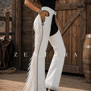 May include: White leather chaps featuring long fringe along the outer legs, a buckled waist belt, and silver-toned metal concho accents. The word ZENTHRA is printed in white capital letters across the center of the image over the white chaps.