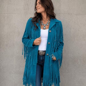 Puede incluir: Una chaqueta de ante color azul cerceta con largos flecos en las mangas y paneles frontales, cuello y botones plateados. La chaqueta se lleva sobre una camiseta blanca lisa y unos vaqueros azul oscuro con un cinturón decorativo.