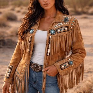 Puede incluir: Una chaqueta de ante color tostado con largos flecos en el pecho y las mangas. La chaqueta está adornada con intrincados abalorios en tonos turquesa, blanco y negro, dispuestos en patrones geométricos en los hombros y los puños.