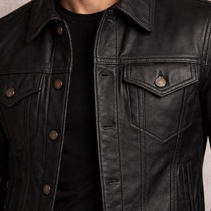 May include: Black leather jacket with a classic collar, button-down front, and two chest pockets. The jacket has a tailored fit and features decorative stitching details. The buttons are a bronze color. A black t-shirt is visible underneath.