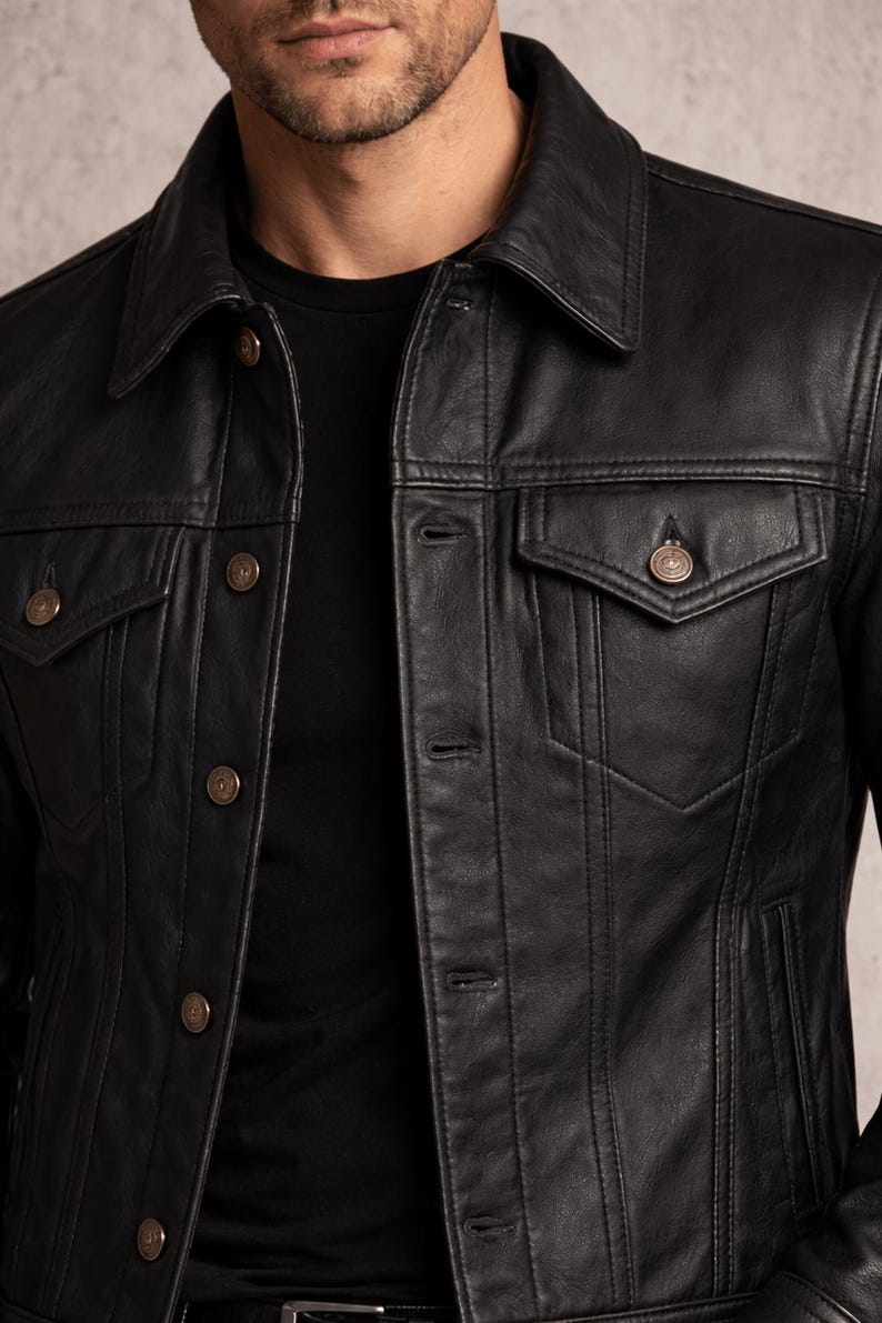 May include: Black leather jacket with a classic collar, button-down front, and two chest pockets. The jacket has a tailored fit and features decorative stitching details. The buttons are a bronze color. A black t-shirt is visible underneath.