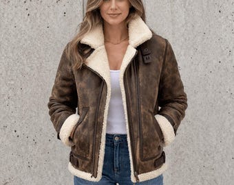 May include: A brown shearling jacket with a cream-colored shearling lining and collar. The jacket features a zip closure, two front pockets, and buckle details. The model is wearing blue jeans and a white tank top.