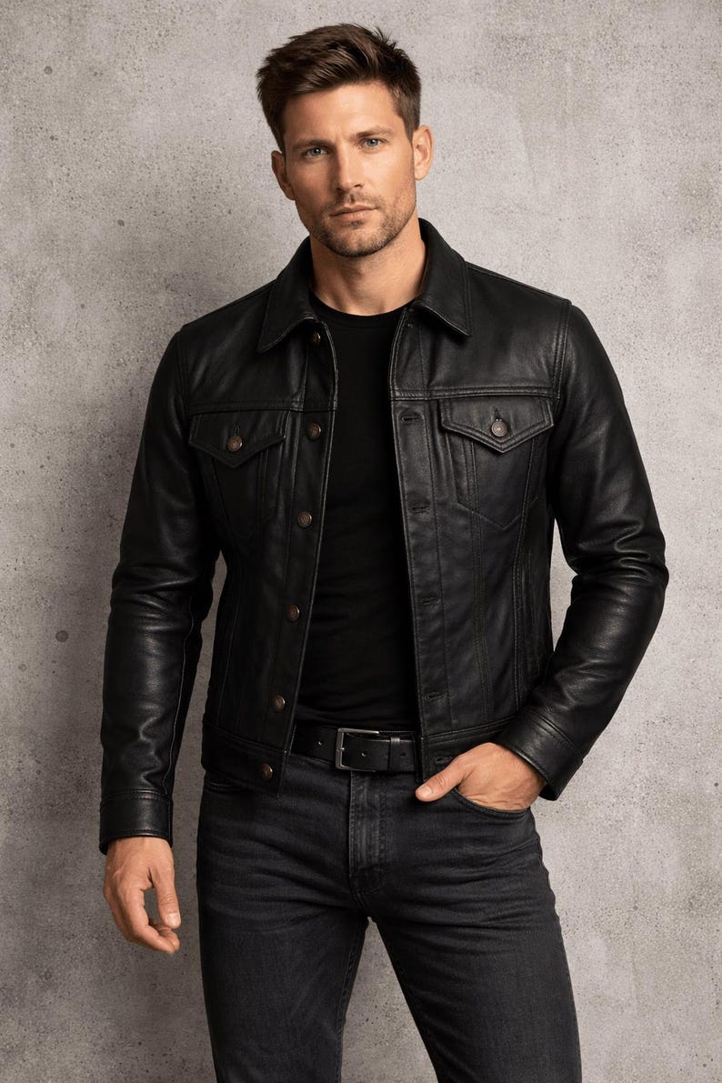 May include: A black leather jacket with button fastenings and two chest pockets. The jacket is worn with a black t-shirt, dark wash jeans, and a black belt. The model is standing in front of a grey background.