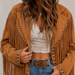 May include: A tan suede jacket featuring long fringe detailing along the sleeves and front panels, accented with silver-toned metal studs lining the lapels and cuffs. The jacket is worn over a white lace crop top and blue denim jeans with a brown belt.