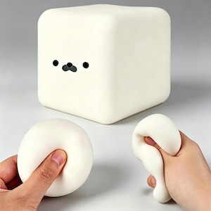 Puede incluir: Un objeto blanco en forma de cubo con una cara de foca de dibujos animados, con ojos y nariz negros. Dos objetos redondos, blancos y blandos están siendo apretados por manos. Los objetos son de un color blanco roto.