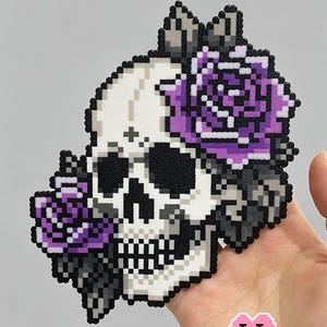 Könnte beinhalten: Pixel-Schädel-Design mit lila Rosen, schwarzen Augen und weißem Hintergrund. Der Text "Purple Rose & Skull" befindet sich oben und das Logo Puff Bead + Patterns unten. Das Design wird von einer Hand gehalten.