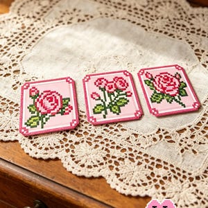 Puede incluir: Tres parches cuadrados rosas con diseños de rosas pixeladas y hojas verdes se muestran sobre un mantel de encaje. Los parches tienen un borde rosa. El texto "Whispers of Roses" y "Puff Bead + Patterns+" son visibles.