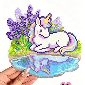 May include: A pixel art craft piece titled Unicorn Amid Floral Brook featuring a white unicorn with a yellow horn and multicolored mane resting beside a blue stream. The scene includes purple lavender flowers and green foliage, with a reflection of the unicorn visible in the water below.