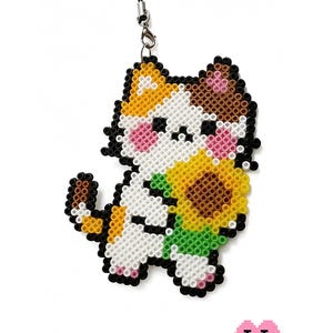 May include: A cat-shaped charm made of fused plastic beads, featuring a white, orange, and brown calico cat holding a yellow sunflower with a green stem. The design includes pink cheeks and black eyes, attached to a black cord with a metal clasp.