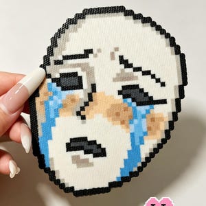 May include: A pixel art bead creation depicting a face with a distressed expression, featuring white skin, black eyebrows, dark eyes, and blue tear streaks running down the cheeks. The design is outlined in black beads on a white background.