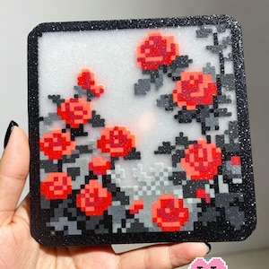 May include: A square coaster with a black glitter border and a white glitter centre featuring a pixel art design of red roses with dark grey leaves and stems. The text Garden of Red Roses is printed at the top and Puff Bead Patterns at the bottom.