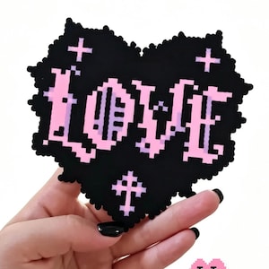 May include: A black heart-shaped pixel art patch featuring the word LOVE in a light pink gradient. The design includes small cross symbols above and below the text. The edges of the heart have a jagged, pixelated texture, creating a distinct digital aesthetic.