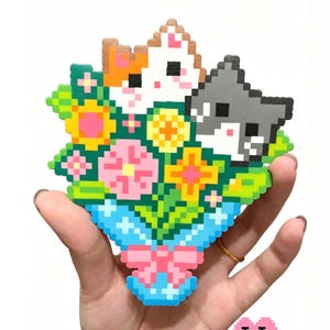 Puede incluir: Un colorido ramo de arte pixelado con tres caras de gato en tonos naranja, blanco y gris, rodeado de flores y vegetación. El ramo se sostiene en un jarrón azul con un lazo rosa. El texto "Cat Bouquet" está en la parte superior.