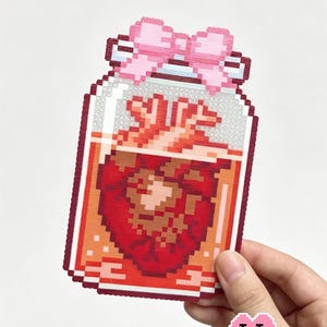 May include: A pixel art style patch featuring a glass jar filled with orange liquid and a red anatomical heart. The jar is topped with a light pink bow. The text Heart Specimen is visible at the top, and Puff Bead Patterns is at the bottom.