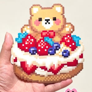 May include: A pixel art sticker titled Strawberry Bear Cake featuring a tan bear with pink cheeks sitting atop a cake decorated with red strawberries, blue blueberries, and white frosting. The design includes a red bow tie on the bear.