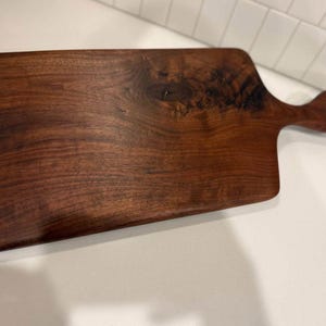 Puede incluir: Una tabla de cortar rectangular de madera con un asa. La tabla es de madera marrón oscuro con un patrón de vetas natural. El asa es curvada y redondeada. La tabla está sobre una superficie blanca.