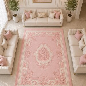 May include: A large, rectangular pink rug with a floral design. The rug features a central floral medallion and a border with repeating floral patterns. The rug is surrounded by cream-colored sofas with pink pillows, creating a cohesive and elegant living room setting.