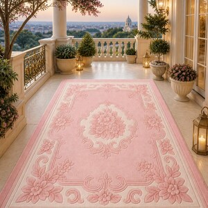 May include: A rectangular pink rug with a floral design. The rug features a central floral medallion and a border with repeating floral and scroll motifs. The rug is placed on a balcony with a view of a cityscape.