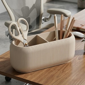 May include: A beige desk organizer with vertical ribbed detailing, holding scissors and pens. The organizer is divided into compartments and sits on a wooden desk. The pens are in shades of beige and brown.