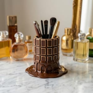 Puede incluir: Un portalápices de cerámica marrón con diseño de tableta de chocolate. El portalápices está lleno de brochas de maquillaje y bolígrafos. La base del portalápices tiene un efecto de chocolate derretido. Frascos de perfume en el fondo.