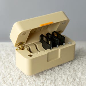 Puede incluir: Un estuche de almacenamiento rectangular de color beige con tapa abatible, que presenta un pequeño detalle naranja en el borde frontal. El interior contiene ranuras moldeadas para tres tarjetas de memoria SD negras y bisagras metálicas doradas.