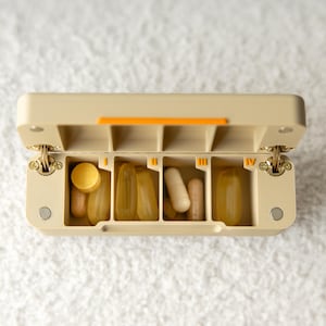 May include: A beige rectangular pill organizer with an open hinged lid. The interior is divided into four compartments labeled with Roman numerals I, II, III, and IV in orange. The compartments contain various yellow, white, and tan capsules.