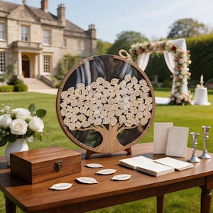 May include: A circular wooden guest book frame featuring a tree silhouette with numerous leaf-shaped wooden inserts inside. The display sits on a wooden table alongside a matching wooden box, stacks of paper cards, and two silver candlesticks.