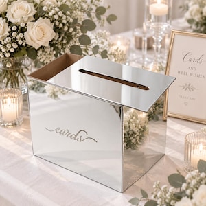 May include: A rectangular mirrored card box with a reflective silver surface and a top slot for inserting items. The front panel features the word cards written in a cursive script font. The box has a clean, modern design with sharp edges.