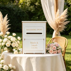Op de afbeelding: Een witte, rechthoekige trouwpostdoos met gouden letters. Het bovenste gedeelte bevat de tekst WEDDING POST boven een horizontale gleuf. Het onderste gedeelte is gegraveerd met de namen Marie & Jakub en de datum 3.10.2026 in een gouden lettertype.
