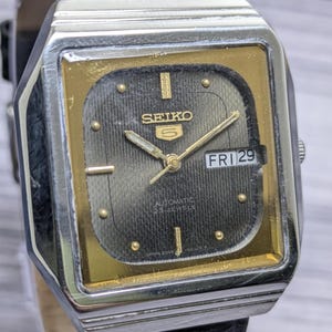May include: A square-shaped Seiko 5 automatic watch with a silver-tone metal case and a black leather strap. The watch face features a dark textured dial with gold-tone hour markers, gold hands, and a day-date window displaying FRI 29.