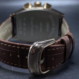 May include: A wristwatch with a brown leather strap featuring a textured pattern and light-colored stitching. The watch has a polished silver-tone metal buckle and a metallic case with visible pushers at the top, viewed from the back side.