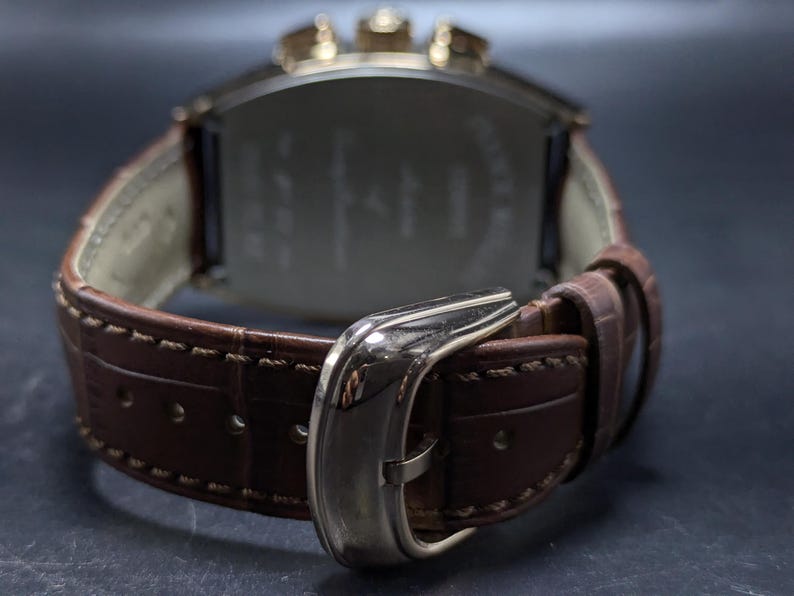 May include: A wristwatch with a brown leather strap featuring a textured pattern and light-colored stitching. The watch has a polished silver-tone metal buckle and a metallic case with visible pushers at the top, viewed from the back side.
