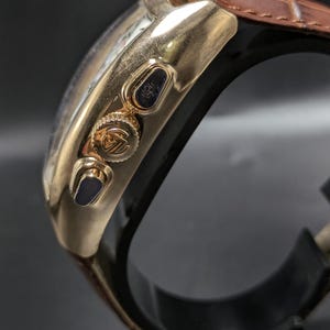 May include: A gold-toned chronograph watch featuring a polished metal case, two side pushers, and a central crown with an embossed logo. The watch is attached to a brown leather strap with a visible textured grain and a black inner lining.