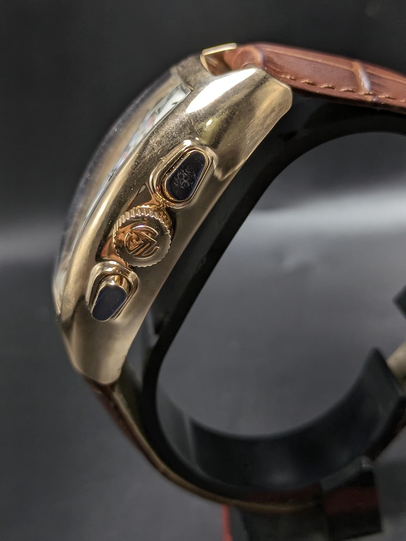 May include: A gold-toned chronograph watch featuring a polished metal case, two side pushers, and a central crown with an embossed logo. The watch is attached to a brown leather strap with a visible textured grain and a black inner lining.